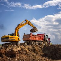 Yellow excavator with engine smoke and empty dump truck working at the construction site.